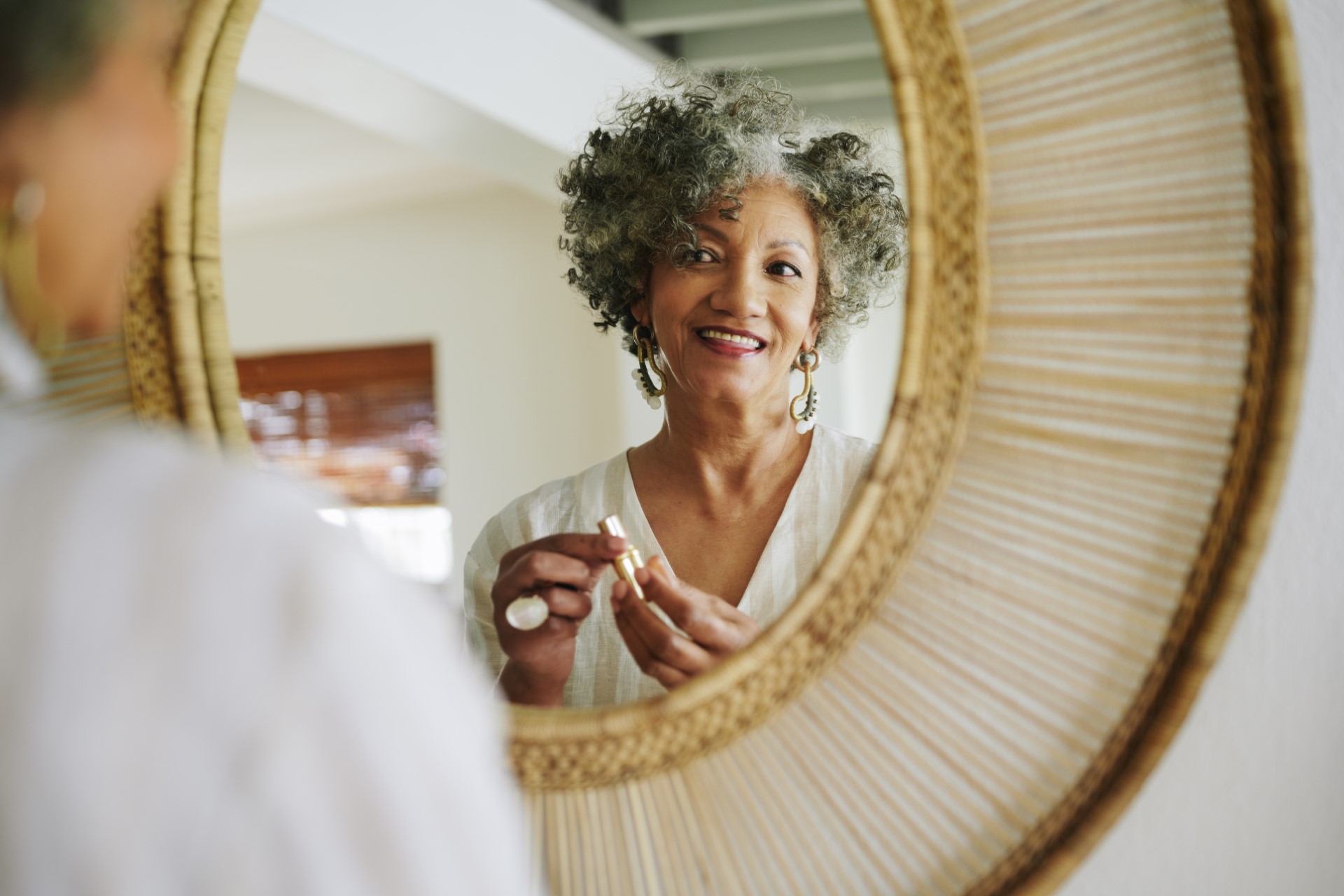 Over the shoulder view of a smiling mature woman putting on lipstick in front of a mirror in a hallway at home